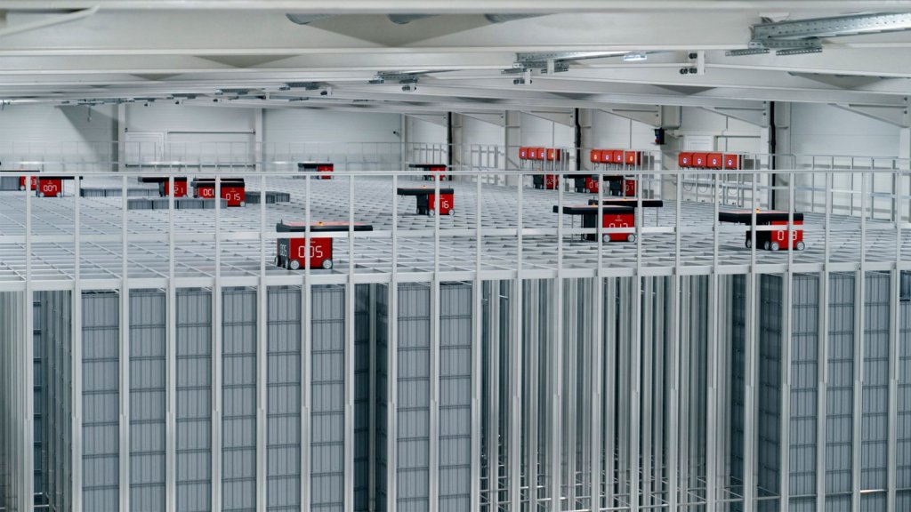 An overhead view of an AutoStore automated warehouse system, showcasing a vast and intricate aluminum cubic grid structure filled with numbered red storage bins. Robots are seen operating on top of the grid, efficiently maneuvering to retrieve and organize the bins for order fulfillment. The high-density storage showcases a modular design, optimized for space-saving and scalable to accommodate various warehouse sizes and needs. The absence of walkways and shelving emphasizes the system's ultra-dense storage capability, designed to enhance operational efficiency, particularly in response to the e-commerce boom and labor challenges. The AutoStore system, paired with sophisticated software integration, underlines a commitment to accelerating picking and replenishment processes, highlighting a future-forward solution to traditional warehouse logistics.