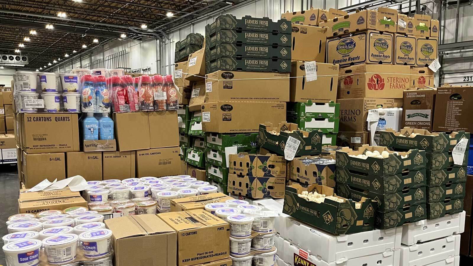 Cases of produce items such as dairy, mushrooms, and lettuce on pallets in a temperature-controlled, cold storage distribution center.