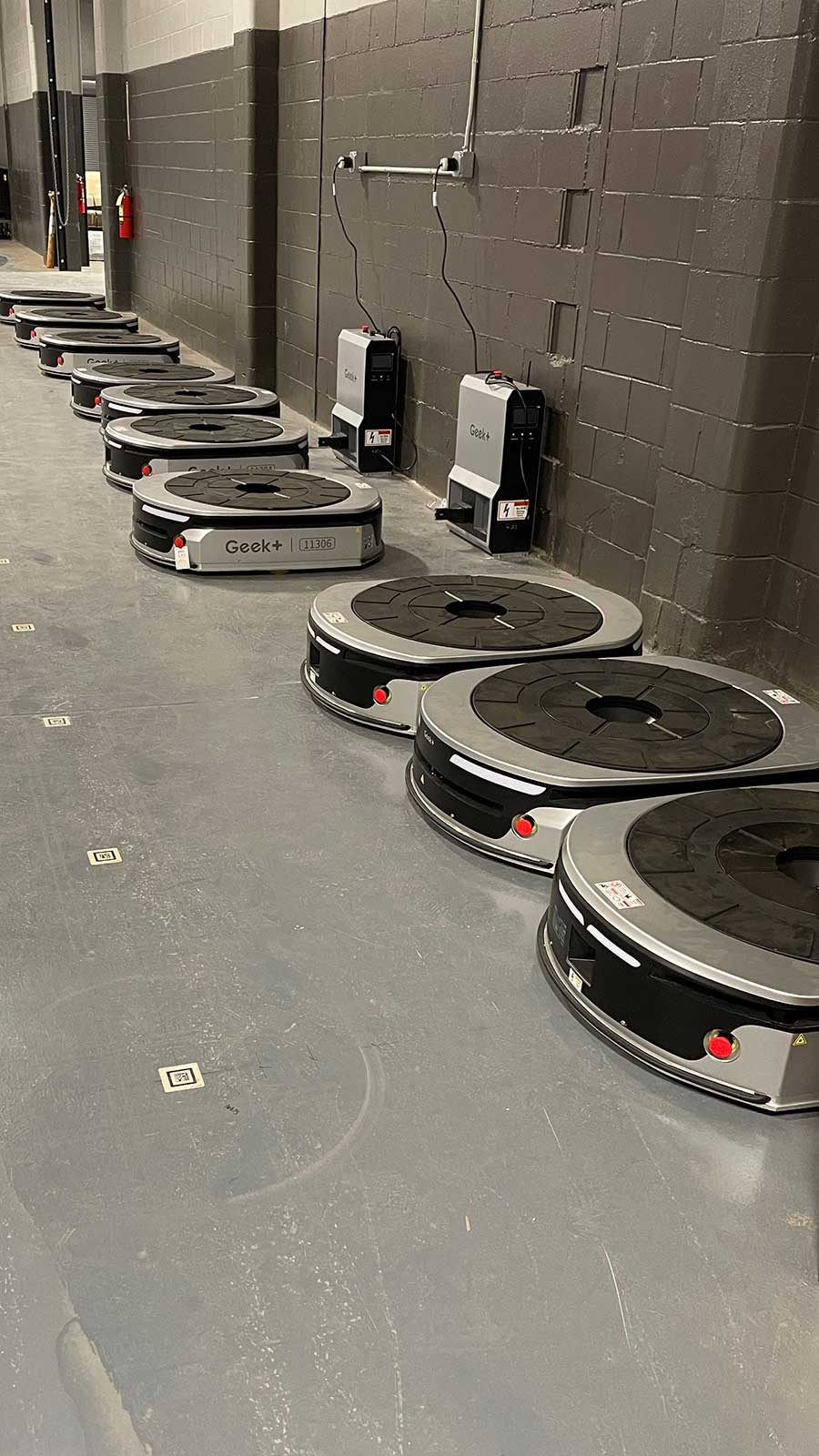 Outdoor Network Case Study Geek+ Integrator. Automated Guided Vehicles (AGVs) lined up against a warehouse wall, showing two charging stations.